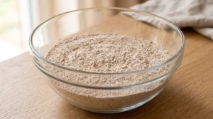 A large glass mixing bowl filled with a well-combined mound of light brown flour mixture and various spices on a wooden counter.