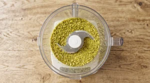 "Toasted pistachios inside a food processor bowl, being ground into fine, vibrant green crumbs."