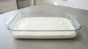 A glass 9x13-inch baking dish on a kitchen counter, showing a smooth, thin layer of white cheesecake filling spread evenly across the bottom.