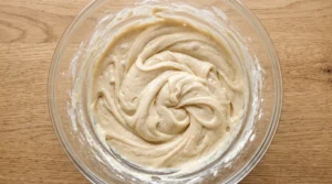 "A large glass bowl with creamy vanilla cake batter, showing the final stage of mixing flour and buttermilk, on a wooden kitchen counter."