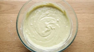 "Pistachio cake batter in a mixing bowl being combined with a dollop of sour cream for a tender texture."