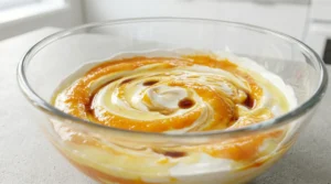 A glass bowl featuring a creamy cheesecake base swirled with vibrant, fresh mango puree, lemon juice, and vanilla extract, ready to be fully combined.