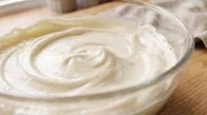 A glass mixing bowl filled with a creamy, speckled vanilla bean frosting, showing a smooth, whipped texture with visible vanilla bean specks, resting on a wooden counter.
