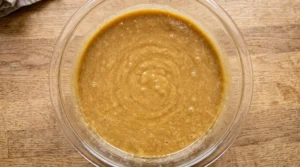 "A large clear glass bowl containing a smooth, golden-brown blended mixture of wet cake ingredients on a light oak wooden kitchen counter."