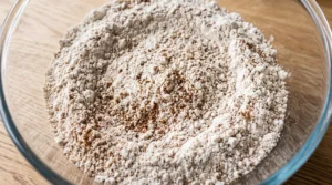 "A large clear glass bowl containing a mixture of flour, spices, and sugar, ready to be whisked together on a light oak kitchen counter."