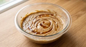 A glass bowl containing cake batter with pure vanilla extract swirled on top on a wooden countertop.