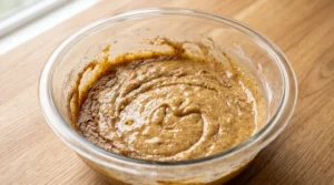 A glass mixing bowl showing dry ingredients being folded into the wet batter on a wooden countertop