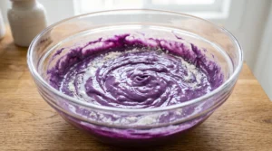 A glass bowl showing flour being mixed into vivid purple ube batter alternately with coconut milk for The Best Ube Cake