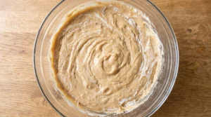 A glass bowl of cake batter blending with alternating additions of flour and buttermilk on a wooden countertop.