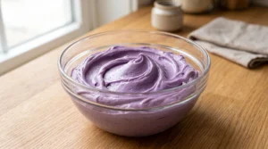 A glass bowl filled with smooth, creamy purple ube buttercream frosting on a wooden countertop.