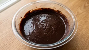 A large glass bowl containing rich, dark chocolate cake batter on a wooden countertop.