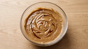A glass bowl containing cookie butter cake batter swirled with vanilla extract on a wooden countertop.