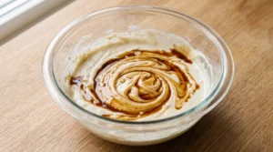 A glass bowl containing cake batter swirled with dark vanilla extract on a wooden countertop.