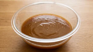 A large glass bowl containing whisked wet ingredients including sugars, oil, applesauce, and reduced cider on a wooden countertop.