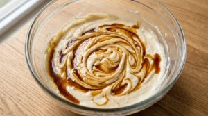 A glass mixing bowl featuring pale cake batter swirled with dark vanilla extract on a wooden countertop.