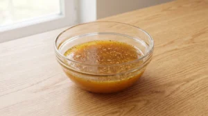 A glass bowl containing melted butter infused with ground ginger, cinnamon, and cloves on a wooden countertop.