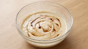 A glass bowl featuring light cake batter swirled with dark vanilla extract on a wooden countertop.