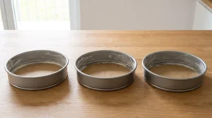 Three greased and floured round cake pans lined with parchment paper on a wooden countertop