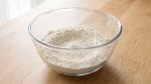 A glass bowl containing flour and other dry ingredients on a wooden countertop.