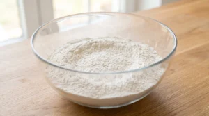 A glass bowl containing measured dry ingredients including cake flour on a wooden countertop.