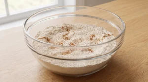 A glass bowl filled with a blend of dry ingredients including cake flour, leavening agents, and spices on a wooden countertop.